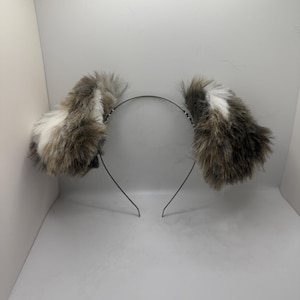 May include: A headband featuring two fluffy faux fur ears. The ears are a blend of brown, grey, and white, secured to a slender, black metal headband. The ears stand upright and have a rounded shape.
