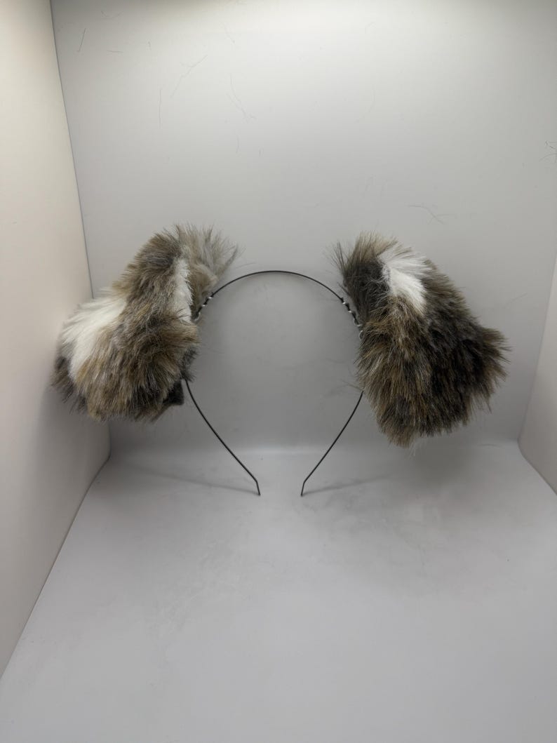 May include: A headband featuring two fluffy faux fur ears. The ears are a blend of brown, grey, and white, secured to a slender, black metal headband. The ears stand upright and have a rounded shape.