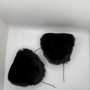 May include: Two black faux fur cat ear headbands. Each ear is a fluffy, triangular shape attached to a thin, flexible wire. The ears are positioned against a white background, creating a simple, clean aesthetic.