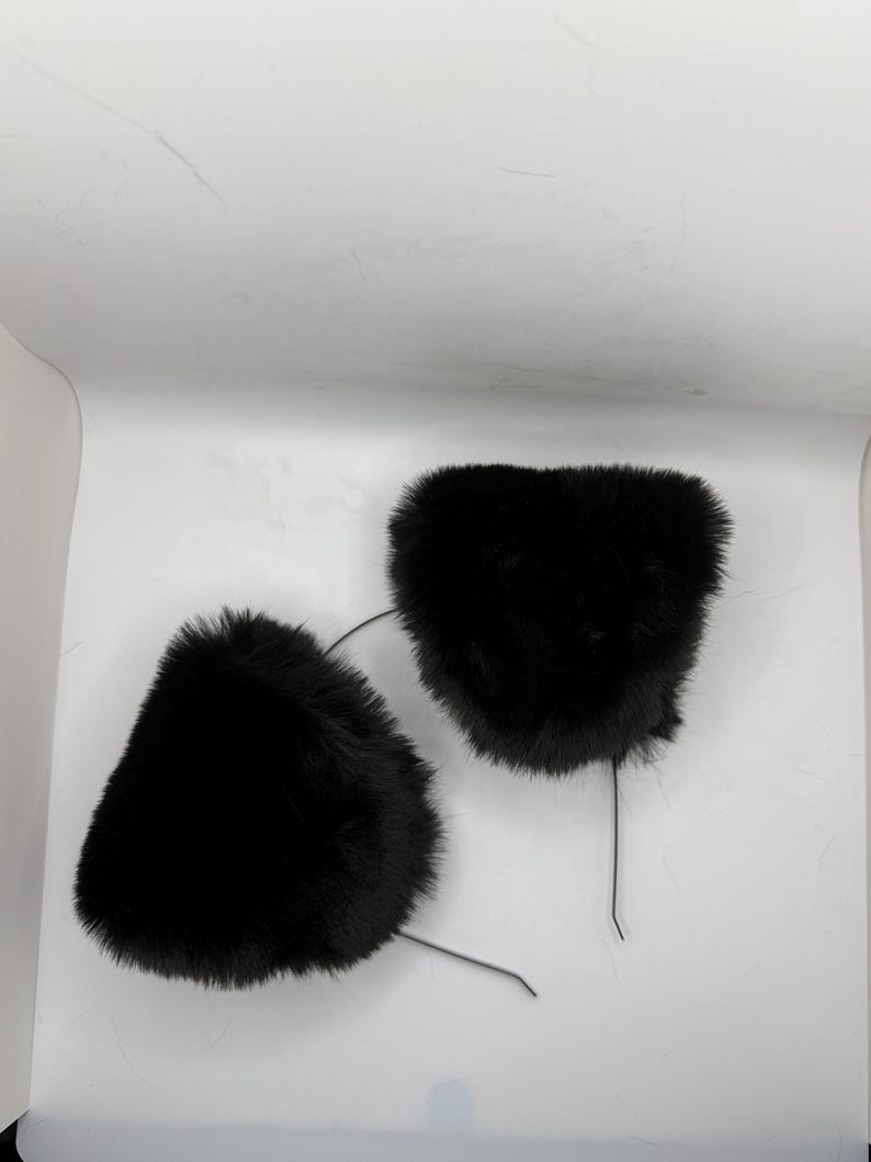 May include: Two black faux fur cat ear headbands. Each ear is a fluffy, triangular shape attached to a thin, flexible wire. The ears are positioned against a white background, creating a simple, clean aesthetic.