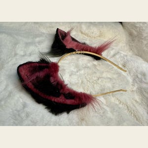 May include: A pair of cat ears on a gold headband. The ears are black and burgundy with pink accents and long, fluffy, pink and black fur. The headband is thin and gold-colored, with a few silver accents.