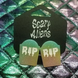 May include: A pair of green and yellow glitter earrings shaped like tombstones with the words "RIP" on them. The earrings are packaged on a black card with the words "Scary Aliens" printed on it.