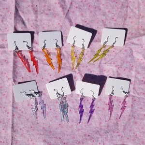 May include: A set of 10 pairs of lightning bolt earrings in various colors, including purple, blue, yellow, orange, and red. The earrings are displayed on white cards on a pink background.