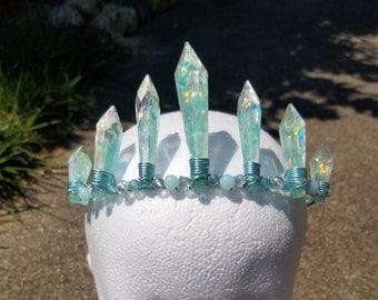 May include: A light blue crystal crown with iridescent accents. The crown is made of wire and beads and features a row of pointed crystals.