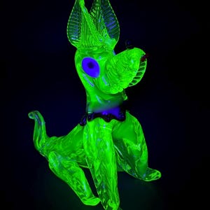 May include: A neon green glass dog figurine with large ears, a purple collar, and a blue eye. The dog is sitting with its tail curled up. The background is black.