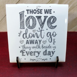 May include: A white ceramic tile with the text "THOSE WE LOVE don't go AWAY they walk beside us Every day" in a black, handwritten style. The tile is displayed on a small wooden stand.