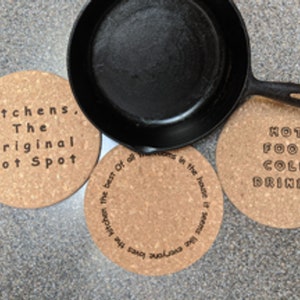 May include: A black cast iron skillet sits on three round cork coasters. The coasters feature text: "Kitchens, The Original Hot Spot" and "Hot Food Cold Drinks". The third coaster has text: "the kitchen the best of all rooms in the house it seems like everyone".