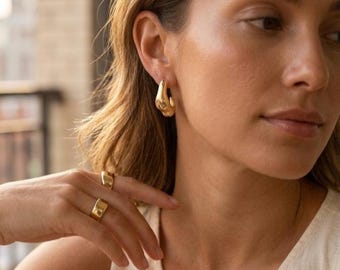 May include: A woman wearing gold-colored jewelry. The jewelry includes a pair of chunky hoop earrings and two rings. The earrings and rings have a smooth, polished finish. The woman is wearing a cream-colored top.
