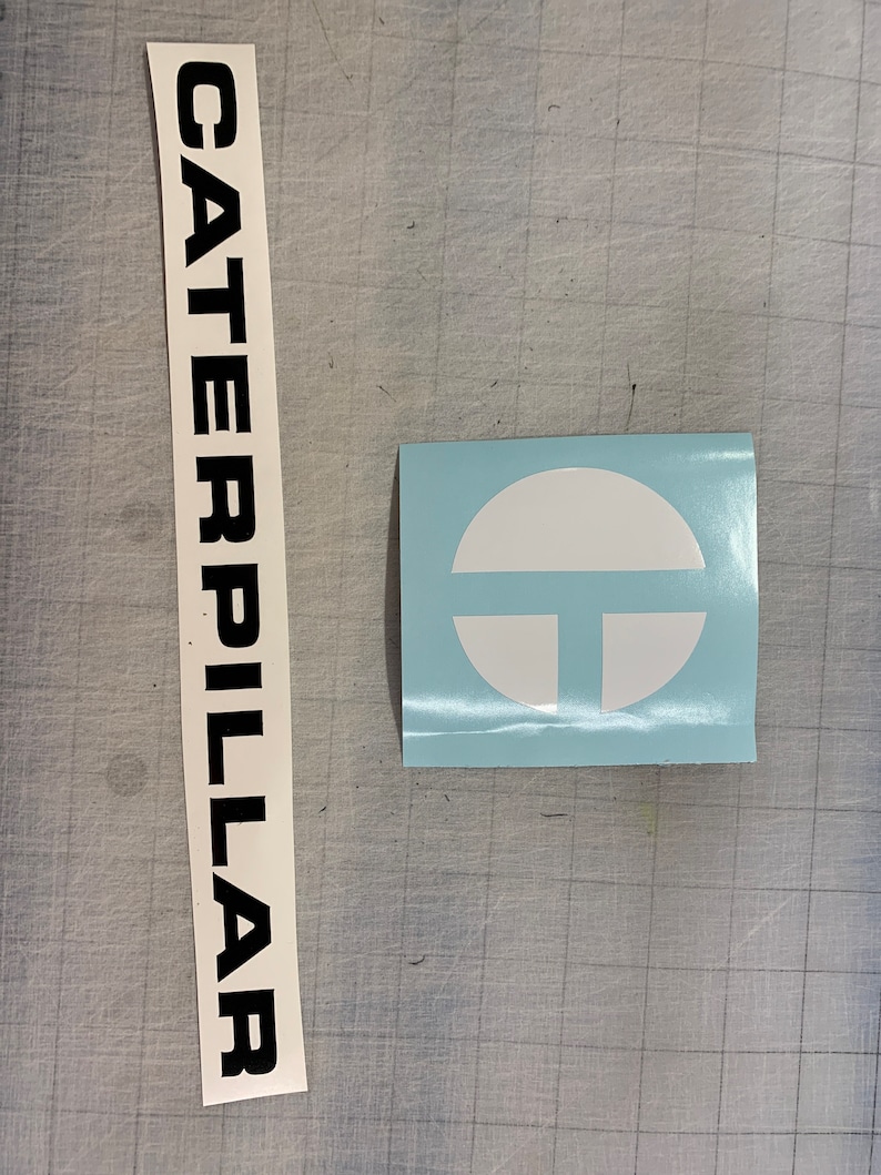 May include: A white sticker with the word "CATERPILLAR" in black lettering and a blue sticker with a white circle containing a stylized letter "T" inside.
