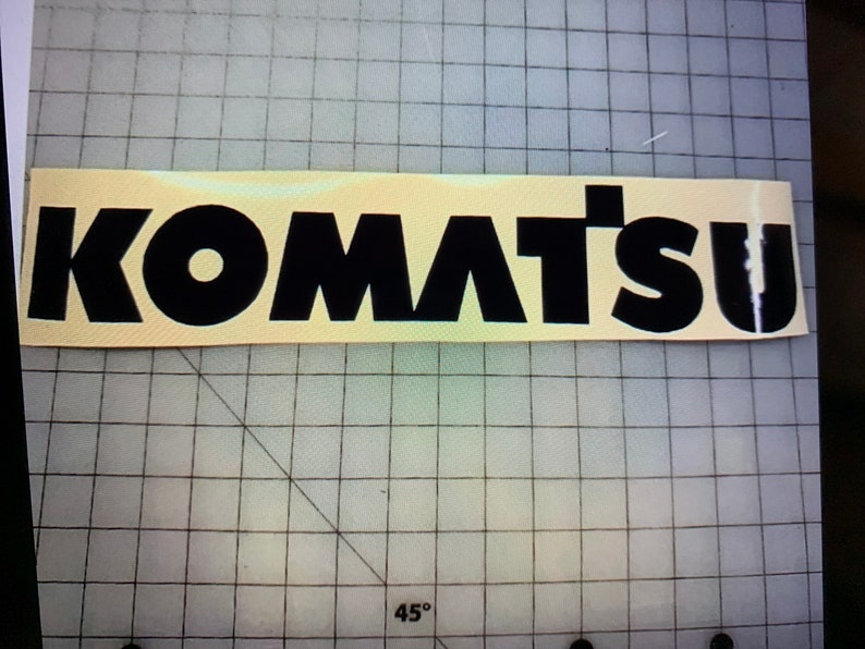 以下が含まれることがあります： 黄色い背景に、太字のブロック体で「KOMATSU」と書かれた黒と白のビニールステッカーです。