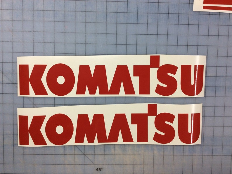 May include: Two white vinyl decals with the word "KOMATSU" in bold, red letters. The decals are on a blue grid background.