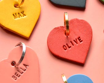 May include: A collection of colorful leather heart-shaped dog tags with personalized names and phone numbers. The tags are attached to gold and silver key rings.