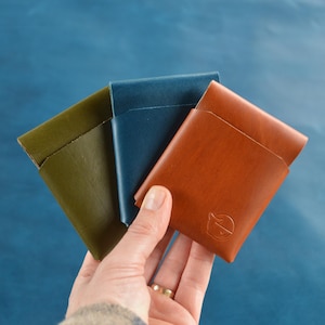 May include: Three leather cardholders in olive green, blue, and brown are displayed against a blue background. The brown cardholder has a logo embossed on the front. The cardholders are held in a hand.