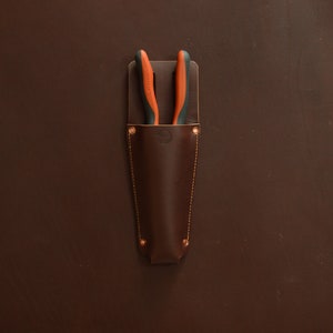 May include: A brown leather tool holder with a pair of orange and black pliers inside.