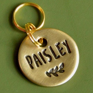 May include: Gold-coloured metal dog tag with the name "Paisley" engraved in black lettering and a small leaf design.