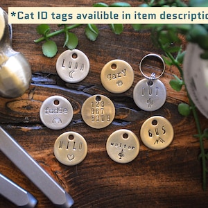K&ouml;nnte beinhalten: Eine Sammlung von neun runden Katzen-ID-Tags aus Metall mit verschiedenen Namen, Zahlen und Symbolen, die darauf graviert sind. Die Tags sind auf einer Holzoberfl&auml;che mit einem Hammer und zwei Metallwerkzeugen angeordnet.