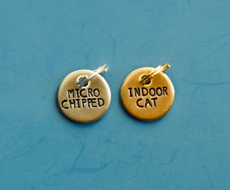 May include: Two small, round pet tags. One is gold-colored and reads "INDOOR CAT" in black lettering. The other is silver-colored and reads "MICRO CHIPPED". Both have a small hole at the top for attaching to a pet's collar.