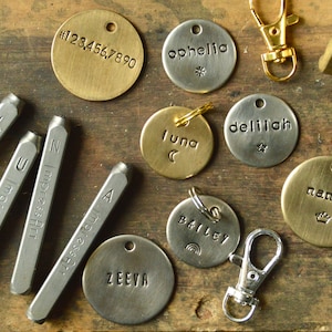 May include: A collection of metal and brass tags with various names and numbers, including "Ophelia", "Luna", "Delilah", "Nando", "Bailey", and "Zeeva".  There are also metal stamps with letters and numbers, a hammer, and a metal key ring.