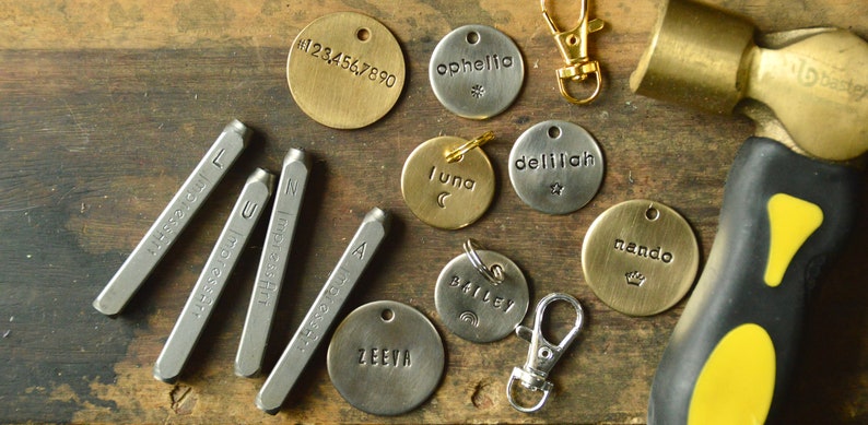 May include: A collection of metal and brass tags with various names and numbers, including "Ophelia", "Luna", "Delilah", "Nando", "Bailey", and "Zeeva".  There are also metal stamps with letters and numbers, a hammer, and a metal key ring.