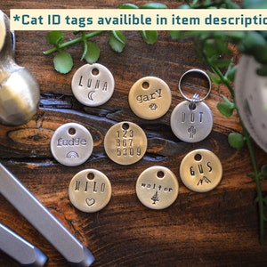 May include: A collection of nine personalized cat ID tags with various designs and names, including Luna, Gary, Dot, Fudge, 123 867 5309, Milo, Walter, and Gus. The tags are made of metal and have a circular shape.