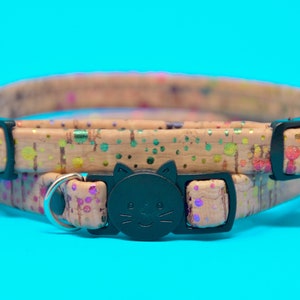 May include: A tan cat collar with a black cat face buckle and a rainbow splatter pattern. The collar has a safety breakaway buckle and a bell.
