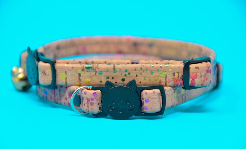 May include: A tan cat collar with a black cat face buckle and a rainbow splatter pattern. The collar has a safety breakaway buckle and a bell.