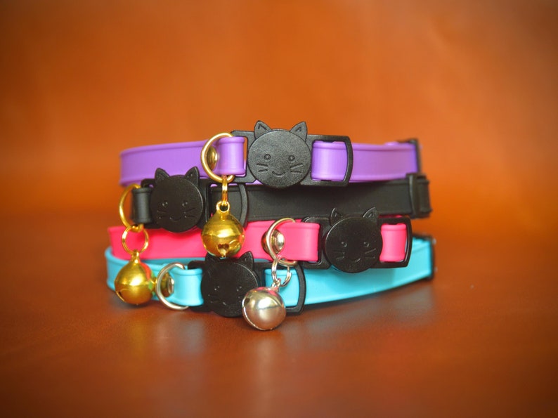 May include: A stack of four colorful cat collars with black cat head charms and gold bells. The collars are purple, black, pink, and blue.