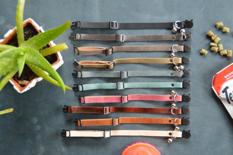 K&ouml;nnte beinhalten: Neun Lederkatzenhalsb&auml;nder in verschiedenen Farben, darunter Grau, Braun, Beige, Rosa und Blau. Jedes Halsband hat einen kleinen schwarzen Katzenanh&auml;nger und eine silberne Glocke.