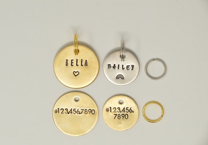 May include: Four round metal pet ID tags with different engravings. Two gold tags read "BELLA" with a heart and "#1234567890". One silver tag reads "BAILEY" with a rainbow. The other gold tag reads "#123.456.7890".