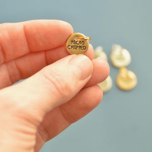 May include: A small, round, gold-colored charm with the words "MICRO CHIPPED" engraved on it. The charm has a small loop at the top. Other charms are blurred in the background.