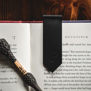 May include: A black leather bookmark with a silver embossed design of a Quidditch player. The bookmark is placed on an open book with the text "QUIDDITCH" visible. The book is open to a page about Quidditch, a magical sport played on broomsticks.