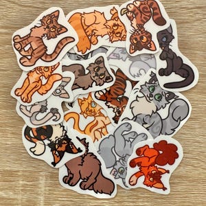Könnte beinhalten: Eine Sammlung von Katzenaufklebern in verschiedenen Farben und Posen. Die Aufkleber zeigen Cartoon-Illustrationen von Katzen in Braun-, Orange-, Grau- und Schwarztönen. Einige Katzen liegen, andere stehen oder spielen. Die Aufkleber befinden sich auf einer hellbraunen Holzoberfläche.
