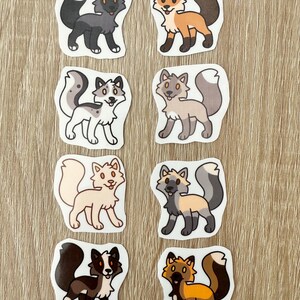 May include: Eight cartoon fox stickers in different colours. The stickers depict foxes in various poses with different fur patterns, including black, orange, white, and grey. Each sticker is outlined in black.