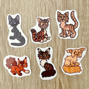 Op de afbeelding: Een verzameling van zes kattenstickers in verschillende poses en kleuren. De stickers tonen cartoon-achtige illustraties van katten in tinten grijs, bruin, oranje en bruin. Elke sticker heeft een witte rand en staat tegen een lichte houtnerf achtergrond.