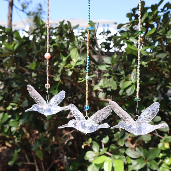 Puede incluir: Tres móviles de viento con forma de colibrí transparente colgando de hilos de cuentas. Los colibríes tienen las alas extendidas y están suspendidos frente a un follaje verde. Las cuentas son en tonos rosa, azul y verde.
