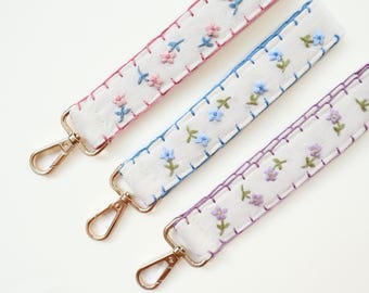 May include: Three white fabric wristlet straps featuring embroidered floral patterns in pink, blue, and purple. Each strap is finished with a decorative blanket stitch border in a matching thread color and includes a gold-toned metal lobster clasp.