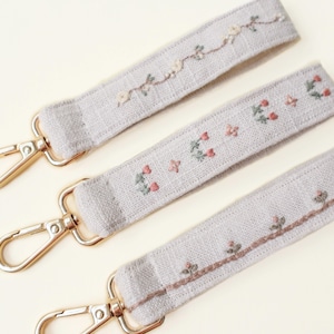 May include: Three pale grey keychains with gold-coloured clasps. Each strap is embroidered with a different floral design in pink, green, and brown. The keychains are made of a textured fabric.