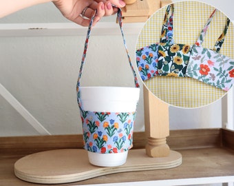 May include: A white to-go cup with a floral print fabric sleeve and a fabric handle. The cup is sitting on a wooden surface. Three different floral print fabric sleeves are shown in the background.