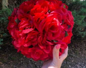 May include: A red rose ball made of artificial flowers. The ball is hanging from a string and is being held by a hand.