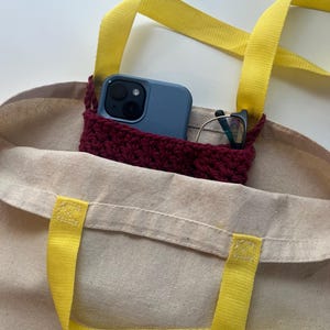 May include: A beige canvas tote bag with bright yellow handles. Inside, a burgundy crocheted pocket holds a blue smartphone and a pair of eyeglasses. The bag is designed for carrying everyday essentials.