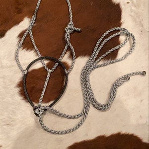 May include: A gray and black rope horse halter with a black noseband and a matching lead rope. The halter is made of braided rope and is designed for use with horses. The background is a brown and white cowhide pattern.
