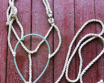 May include: A white rope halter with a green rope noseband. The halter is laid out on a red wooden surface.