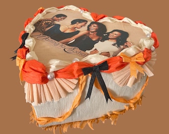 Könnte beinhalten: Eine herzförmige dekorative Schachtel mit einem gedruckten Bild von vier Frauen und dem Text "Waiting to Exhale". Die Schachtel ist mit orangefarbenem und cremefarbenem Papier, Schleifen und Perlen verziert.