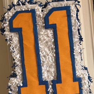 May include: A number 11 piñata featuring a bright orange center outlined in dark blue, surrounded by a thick border of white fringed paper with scattered dark blue fringe accents along the outer edges of the numerical shape.