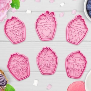 May include: Six pink plastic cupcake cookie cutters with different designs. The cutters are on a white wooden surface.
