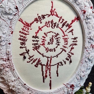 Op de afbeelding: Een wit ovaal frame met sierlijke details omringt een kruissteekontwerp. Het ontwerp heeft een rood en wit bloemenpatroon met het woord "Violet" in het midden. Rode kralen zijn rond het ontwerp verspreid.