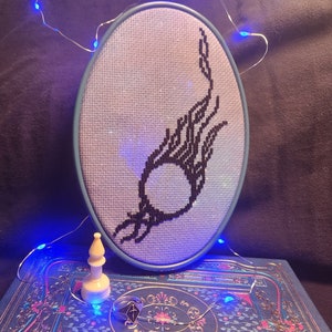 May include: A blue embroidery hoop with a white background and a black embroidered design of a flame with a circle in the center. The hoop is sitting on a blue and gold patterned surface.