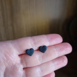 May include: A pair of small, matte black heart-shaped stud earrings. Each earring features a simple, solid heart silhouette with a smooth, uniform finish, designed to be worn as a pair of minimalist ear accessories for everyday use.
