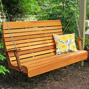 May include: A wooden porch swing with a slatted back and seat, suspended by chains. The swing is made of light brown wood and has armrests. A decorative pillow with yellow flower pattern rests on the seat. The swing is outdoors, surrounded by greenery.