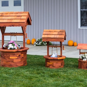 May include: Three wooden well planter boxes in different sizes, with a brown stain finish. The largest well planter has a roof and is filled with pink flowers. The medium-sized well planter is filled with blue flowers. The smallest well planter is filled with white flowers.